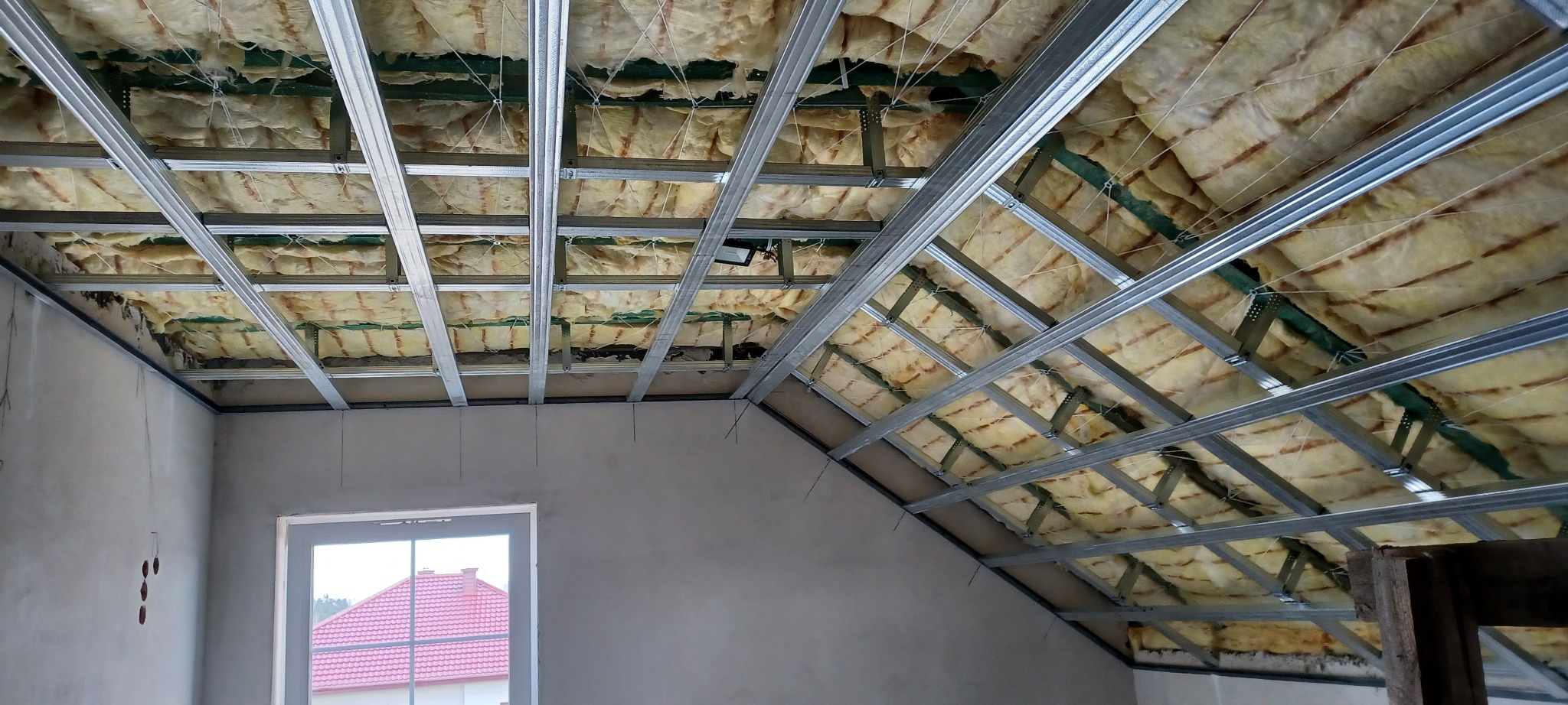 Skośny sufit poddasza z izolacją z wełny mineralnej i metalowym stelażem pod płyty gipsowe, widok z dołu, okno w tle.