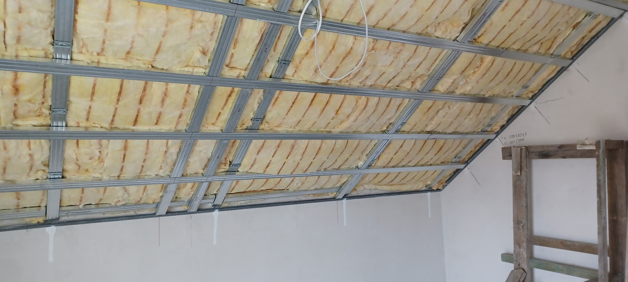 Skośny sufit poddasza z izolacją z wełny mineralnej, widoczne metalowe profile konstrukcyjne. Fragment ściany z odręcznymi notatkami i drewnianą konstrukcją w tle.