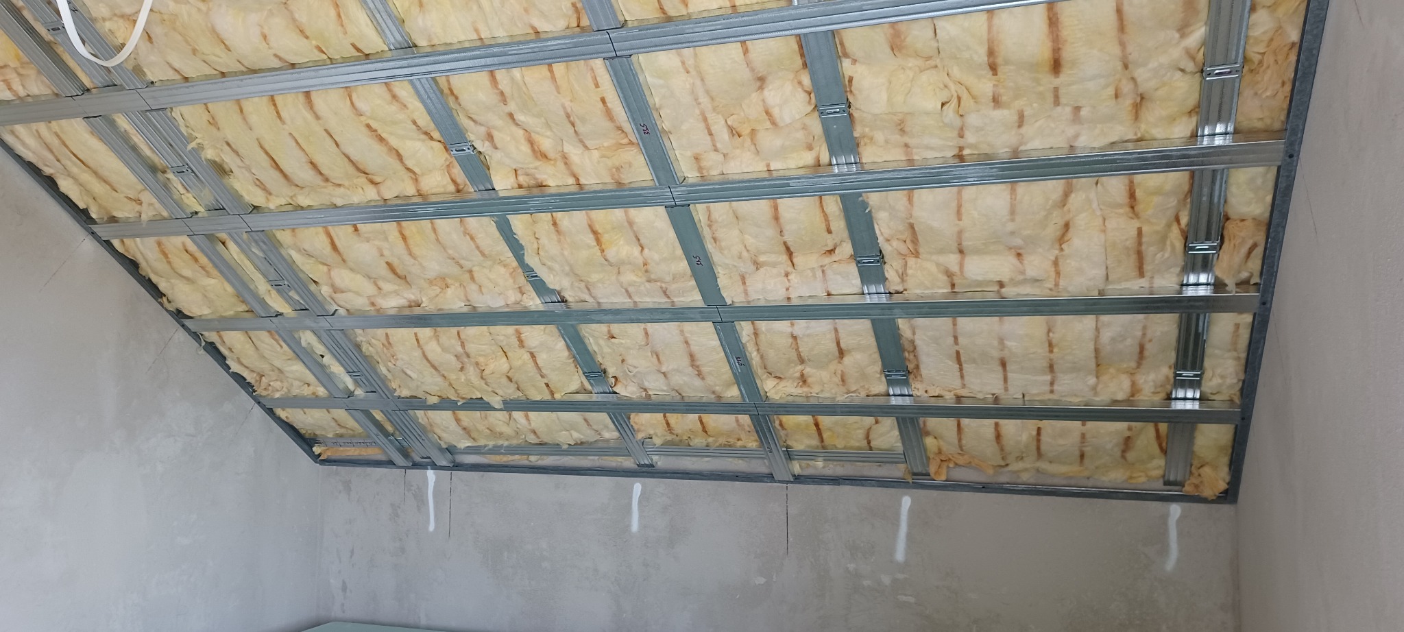 Skośny sufit poddasza z izolacją z wełny mineralnej, widoczną między metalowymi profilami. Szare ściany w tle. Ujęcie z dołu, ukazujące konstrukcję przed zabudową.