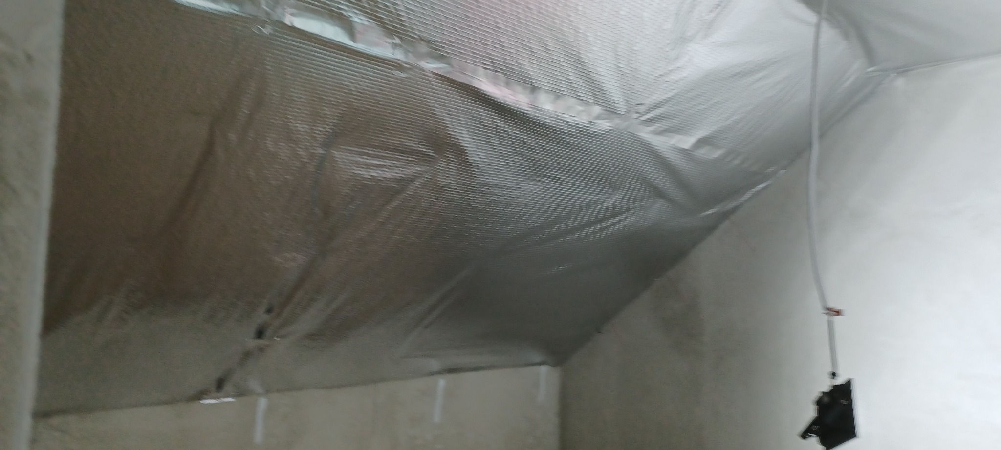 Izolacja termiczna poddasza z folią aluminiową, widoczne łączenia i fragment ściany. Oświetlenie górne, surowy stan wykończenia.