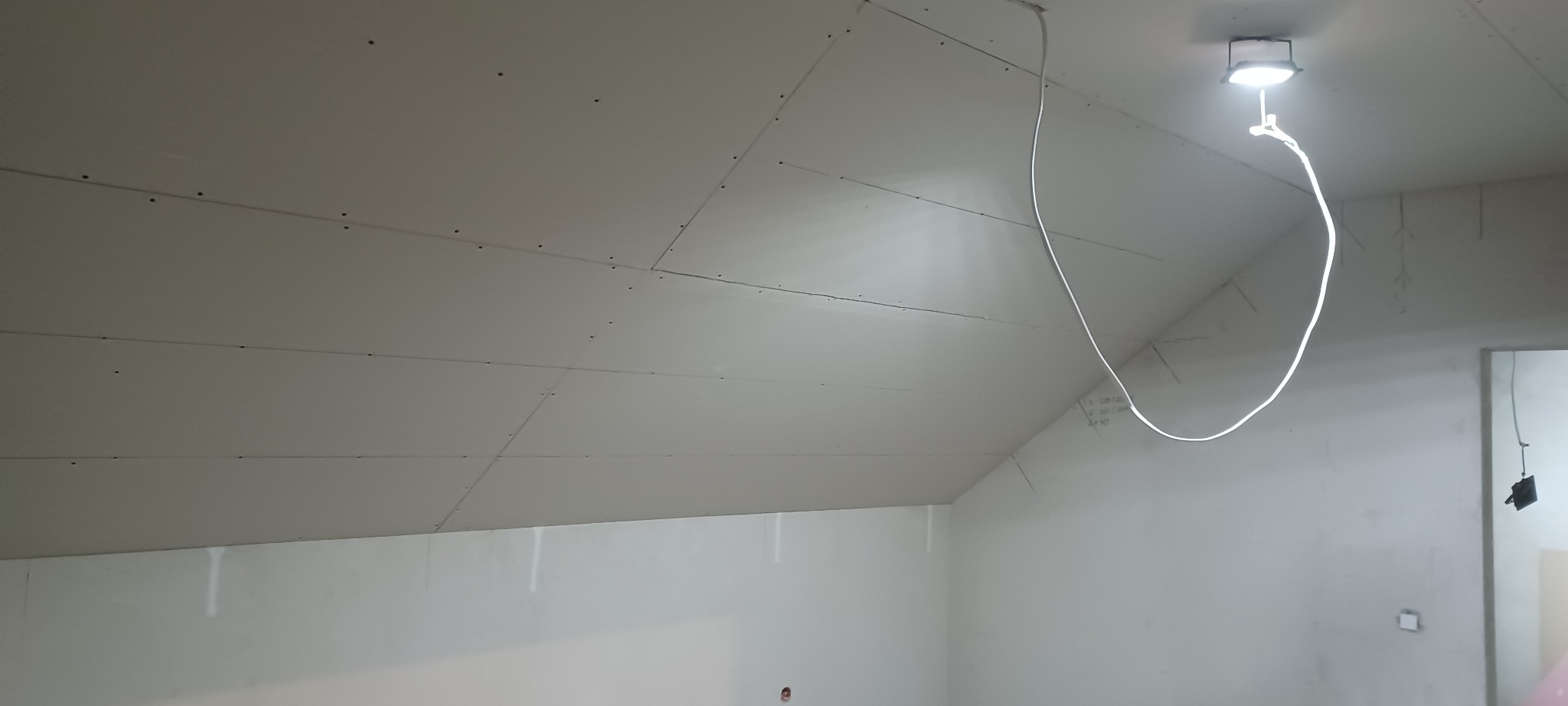 Poddasze z zamontowanymi płytami kartonowo-gipsowymi, widoczne łączenia i śruby, oświetlone lampą budowlaną. Skos dachu zarysowany ołówkiem.
