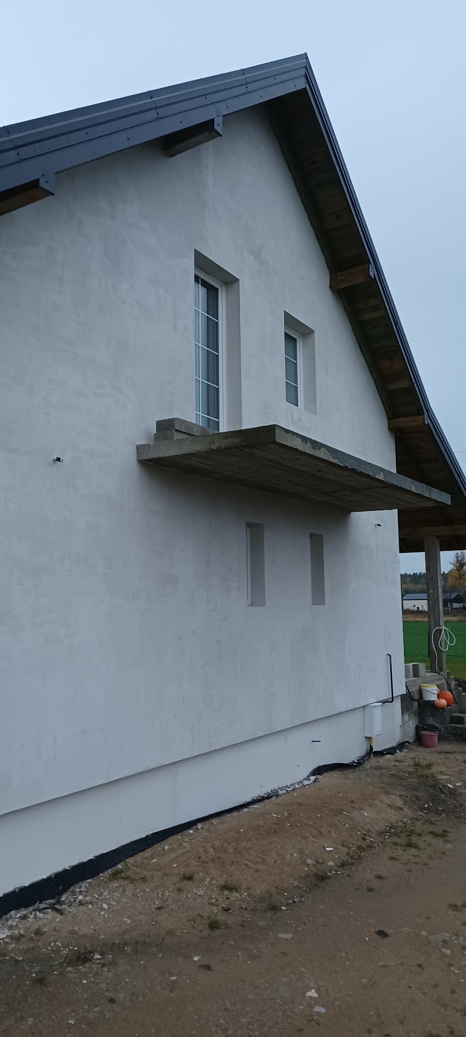 Boczny widok domu z jasną elewacją, widoczne okna, betonowy balkon i fragment dachu. Teren wokół domu częściowo zagospodarowany, w tle zieleń.