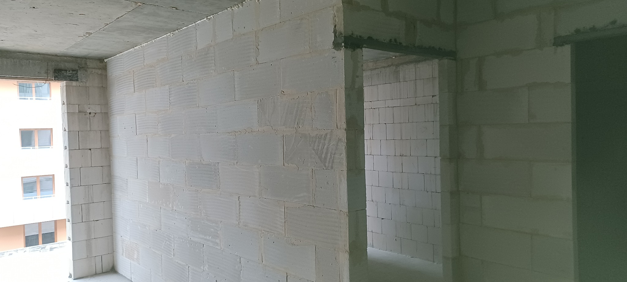 Wnętrze w stanie surowym: ściany z białych bloczków, widoczne okno z budynkiem w tle i otwór drzwiowy. Surowy betonowy strop. Widok z rogu pomieszczenia.