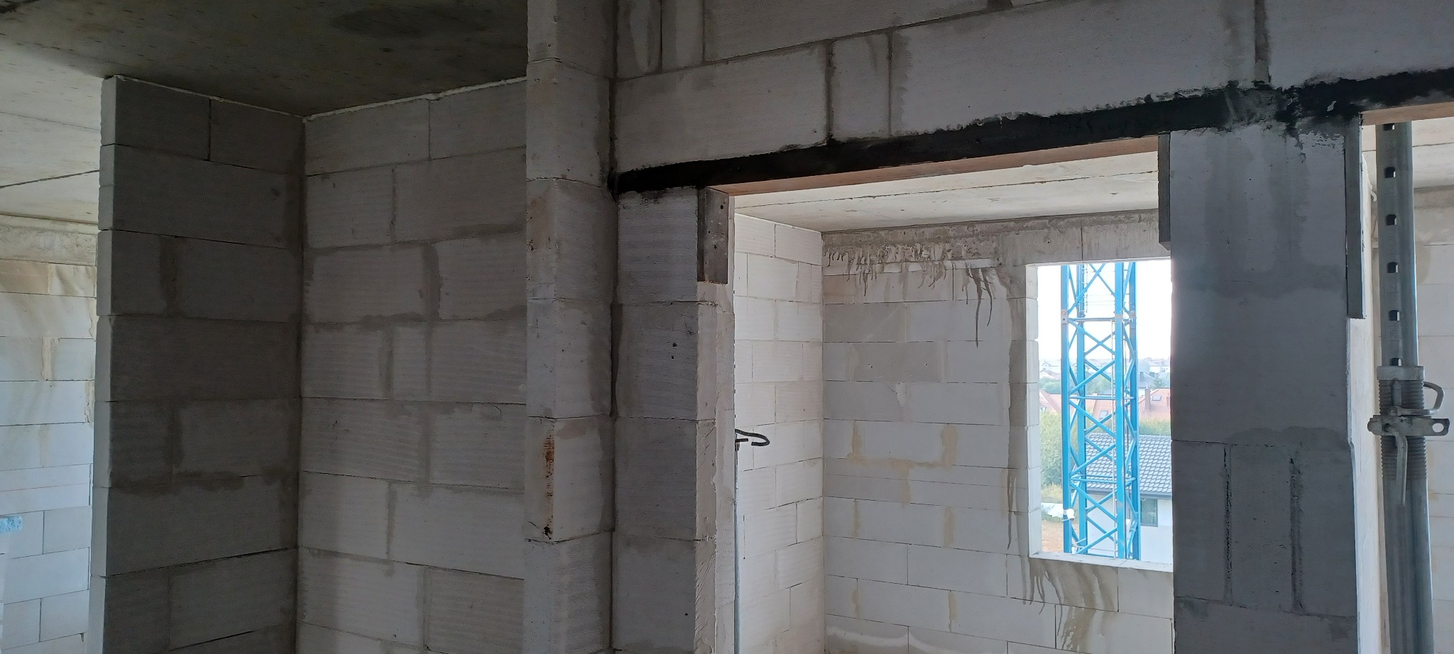 Wnętrze w trakcie budowy: ściany z białych bloczków, widoczne nadproże i okno z widokiem na żuraw budowlany. Surowy stan, przygotowanie do dalszych prac wykończeniowych.