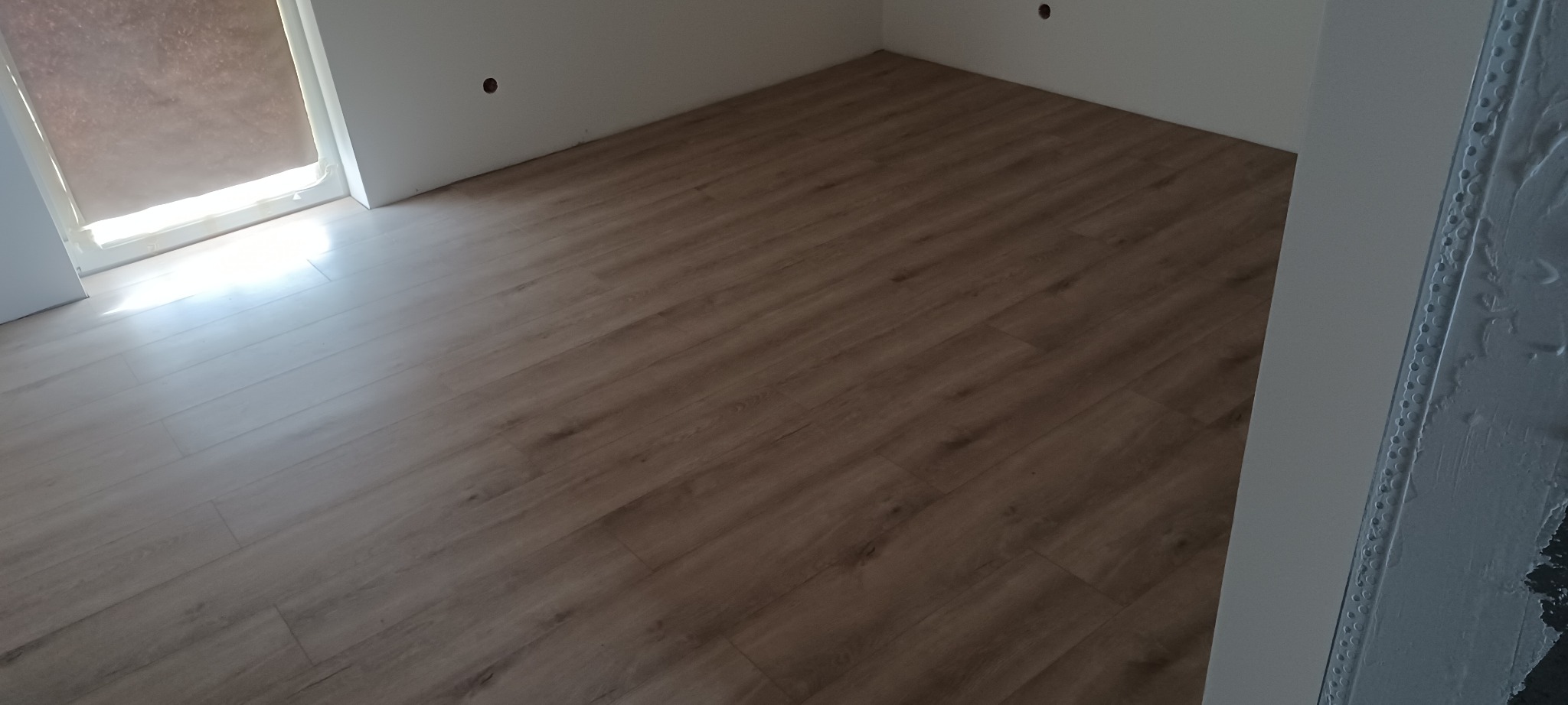 Ukończona podłoga z paneli w odcieniach drewna, widoczna w jasnym pomieszczeniu z białymi ścianami i oknem z roletą. Świeżo wykończone ściany.