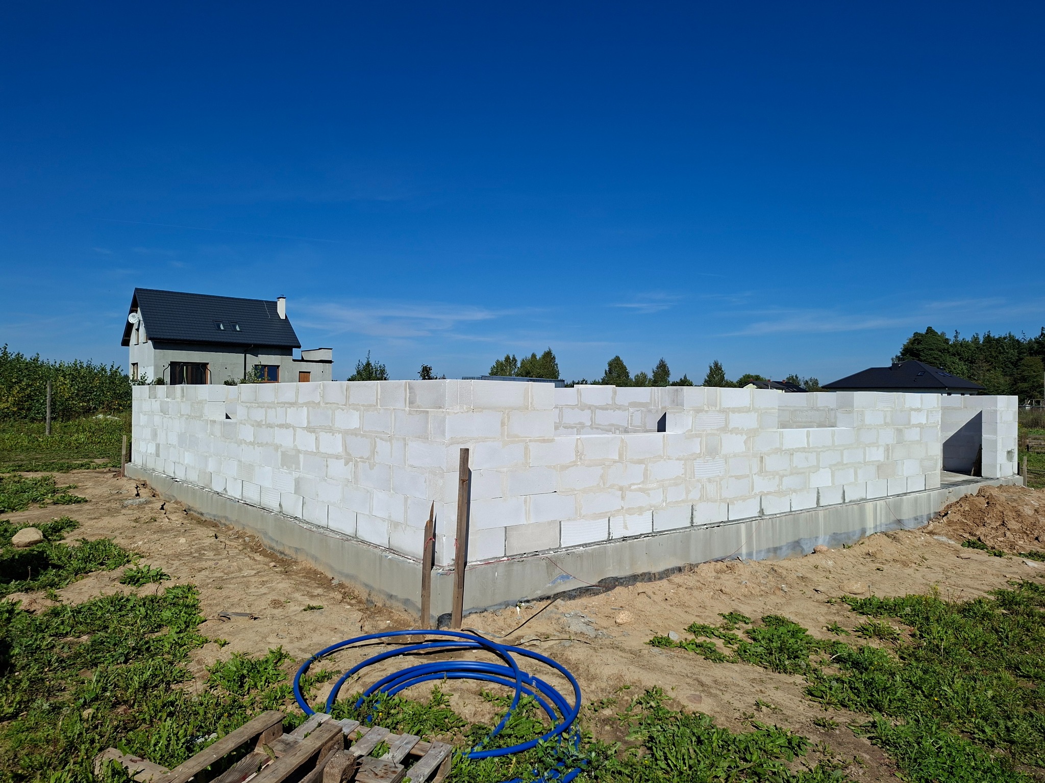 Wznoszenie ścian parteru domu z betonu komórkowego na fundamencie. Widoczne niebieskie rury instalacyjne i dom w tle pod błękitnym niebem.
