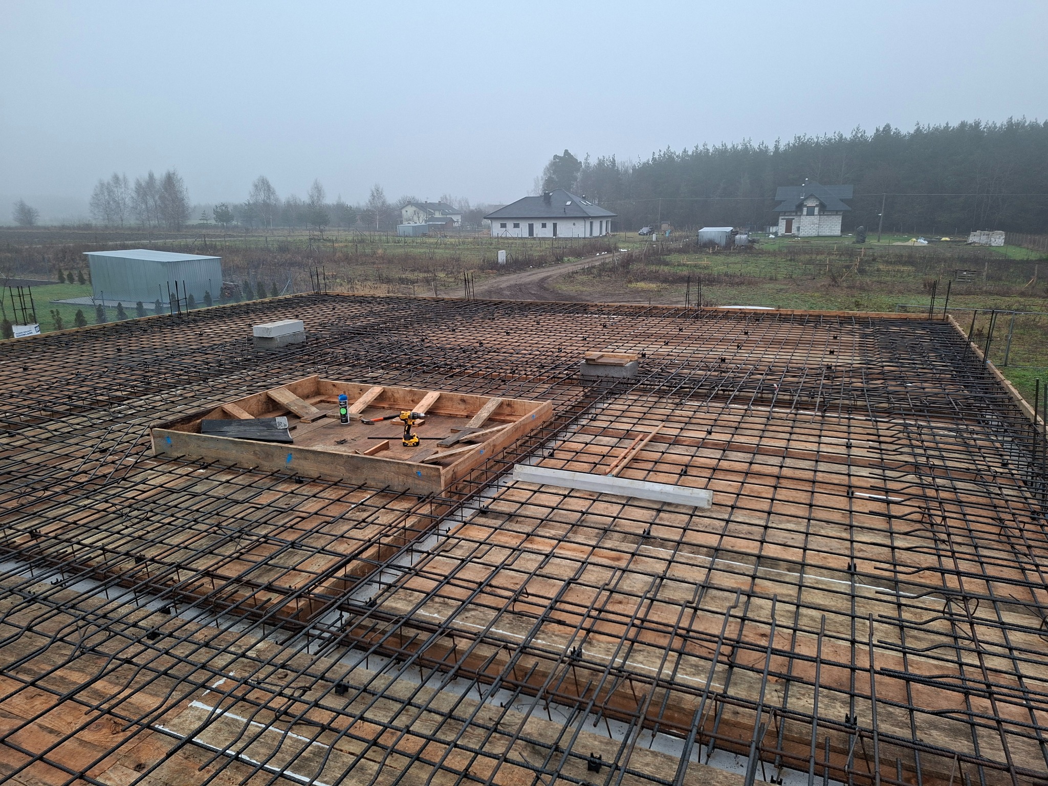 Zbrojenie płyty fundamentowej na drewnianym szalunku, widok z góry. W tle zabudowa jednorodzinna w porannej mgle.
