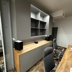 Elżbieta Kutyła Przedsiębiorstwo Wielobranżowe - Szara szafa na wymiar z drewnianym blatem i oświetleniem LED, uzupełniona o czarne głośniki. Dwa krzesła biurowe i biurko z blatem z litego drewna w tle.