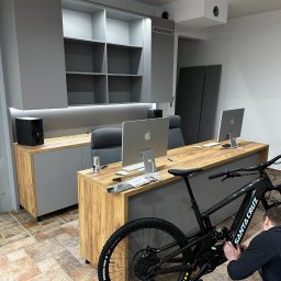 Elżbieta Kutyła Przedsiębiorstwo Wielobranżowe - Nowoczesne biuro z szafami na wymiar w odcieniach szarości i drewna, z rowerem Santa Cruz w kadrze. Osoba reguluje przerzutki. Dwa monitory Apple na biurku.