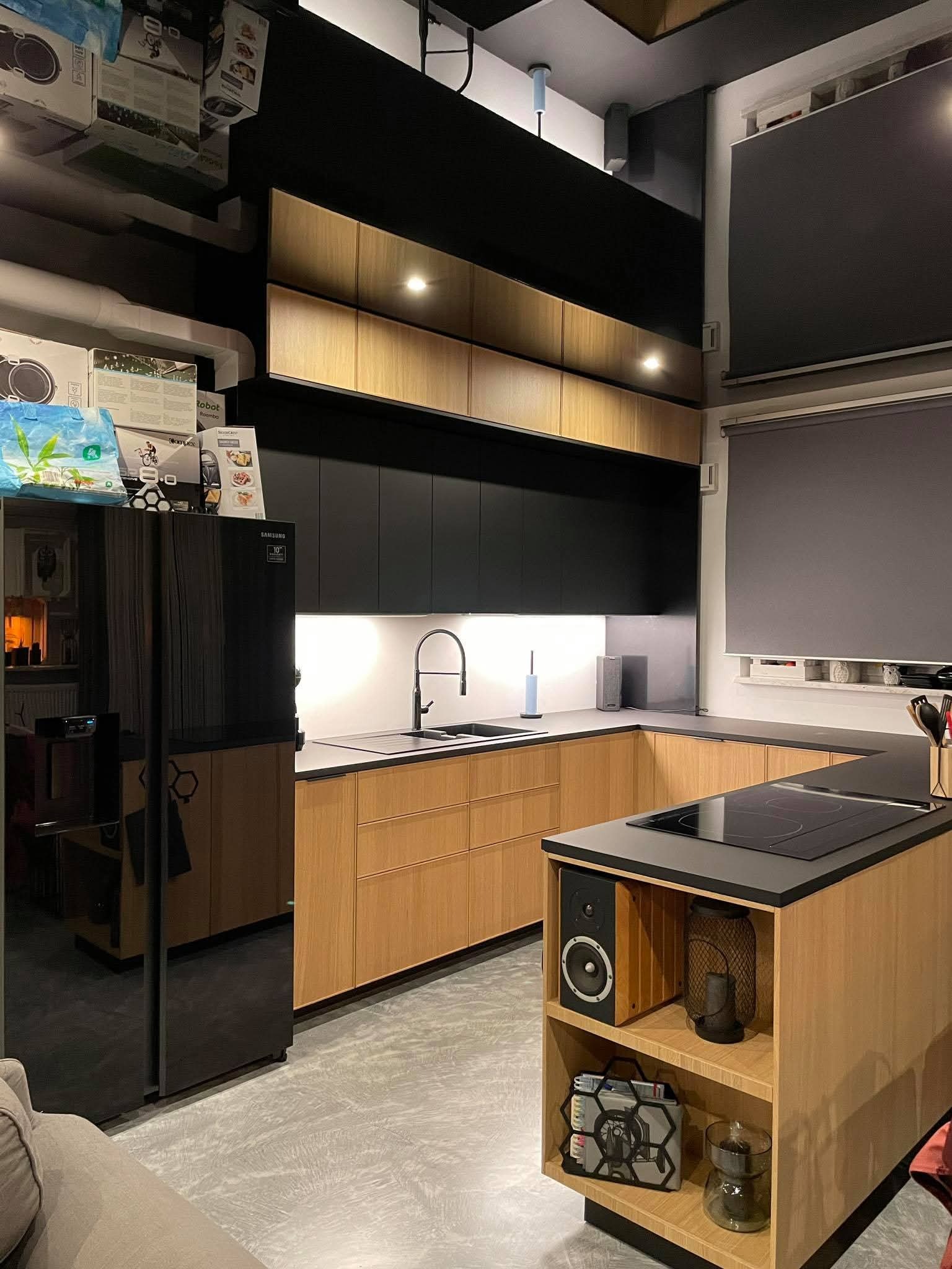 Nowoczesna kuchnia na wymiar z drewnianymi frontami i czarnymi elementami w Pruszkowie. Widoczna wyspa kuchenna z płytą indukcyjną i półkami.