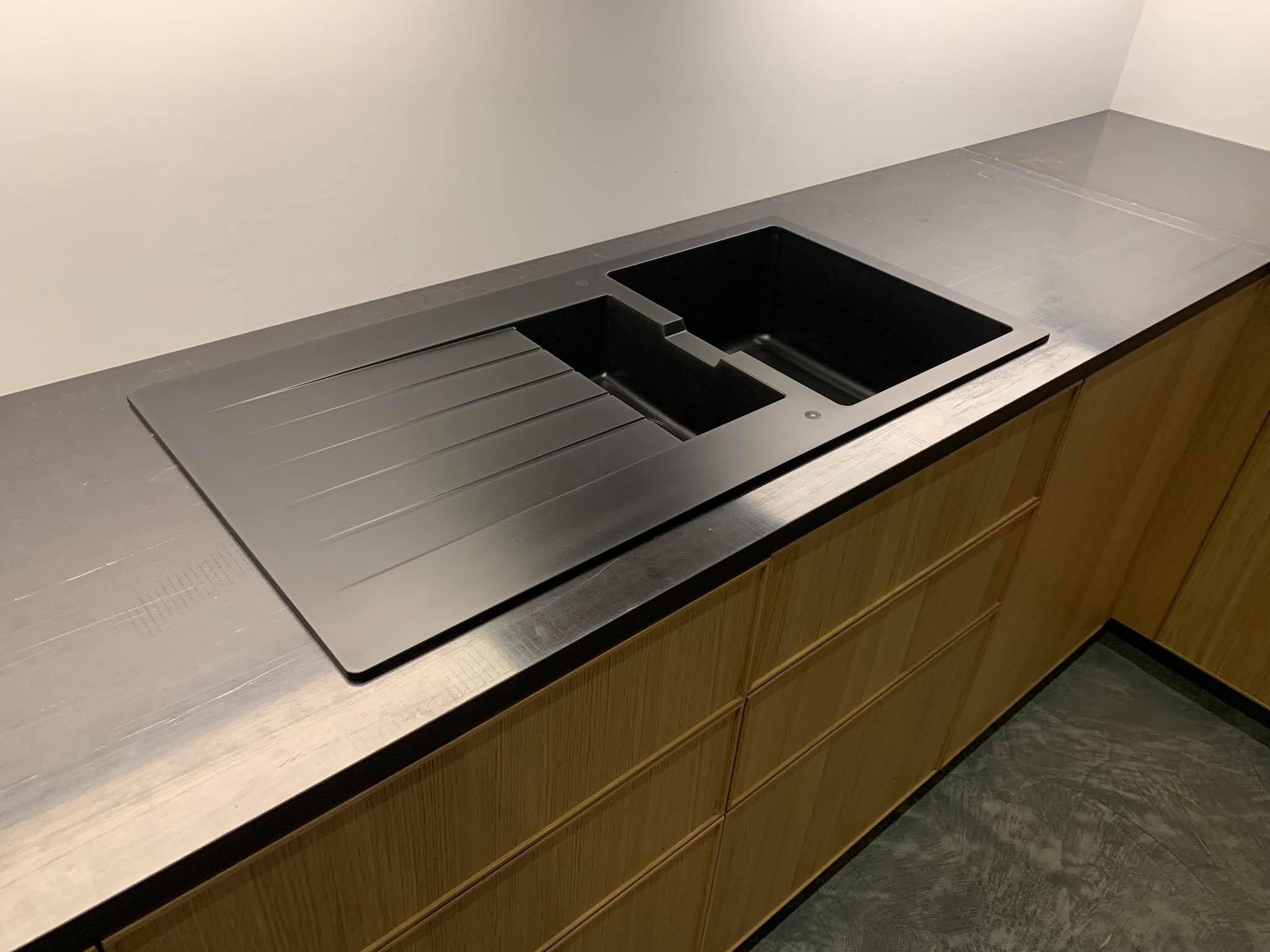 Czarny, nowoczesny zlew granitowy z ociekaczem, wkomponowany w drewniany blat kuchenny. Minimalistyczny design, jasne drewno szafek, szary blat i tło.