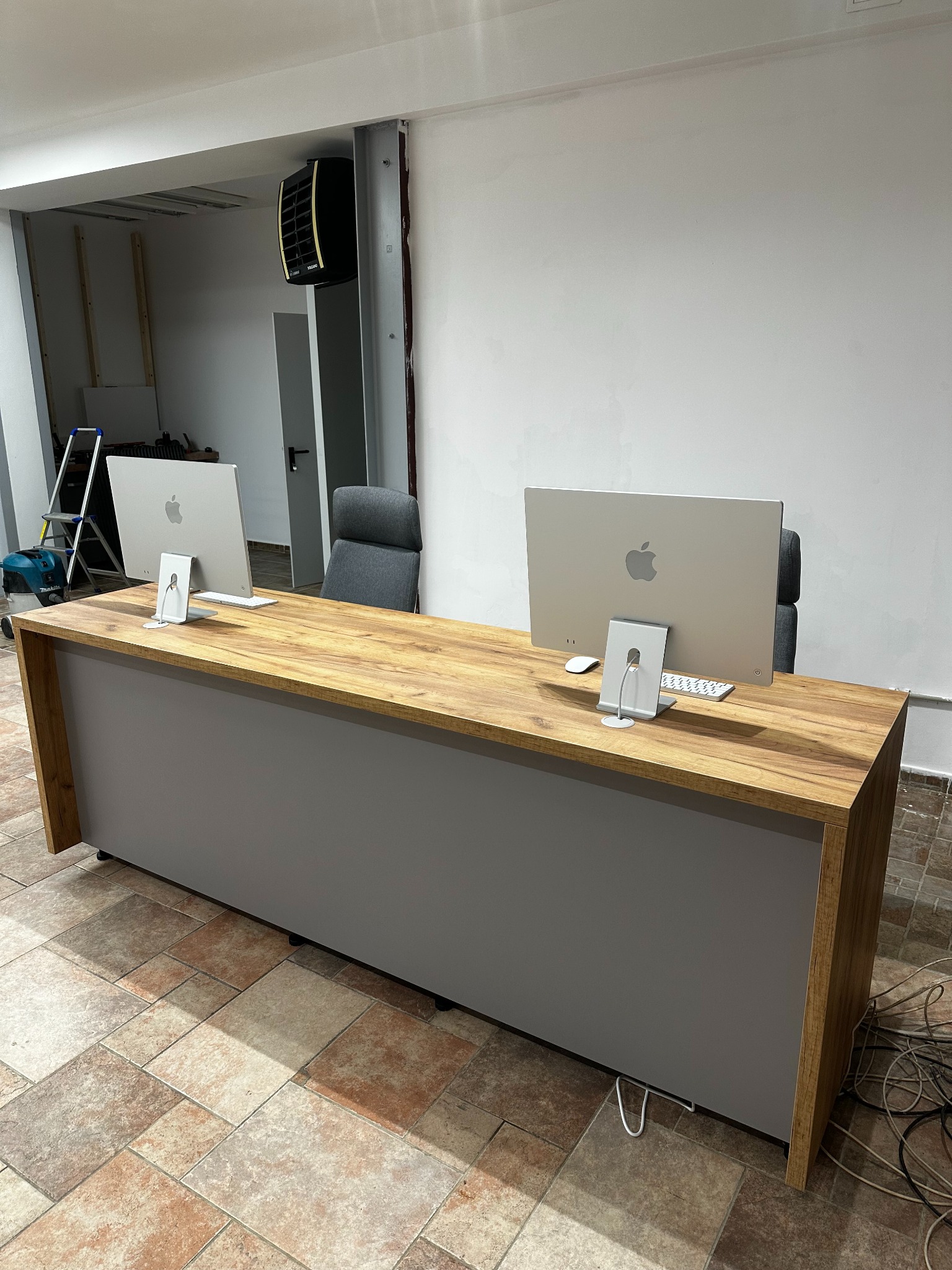 Biurko na wymiar z drewnianym blatem i szarą obudową, dwa komputery Apple, krzesła biurowe w tle, widok z przodu, płytki na podłodze, przewody na podłodze.