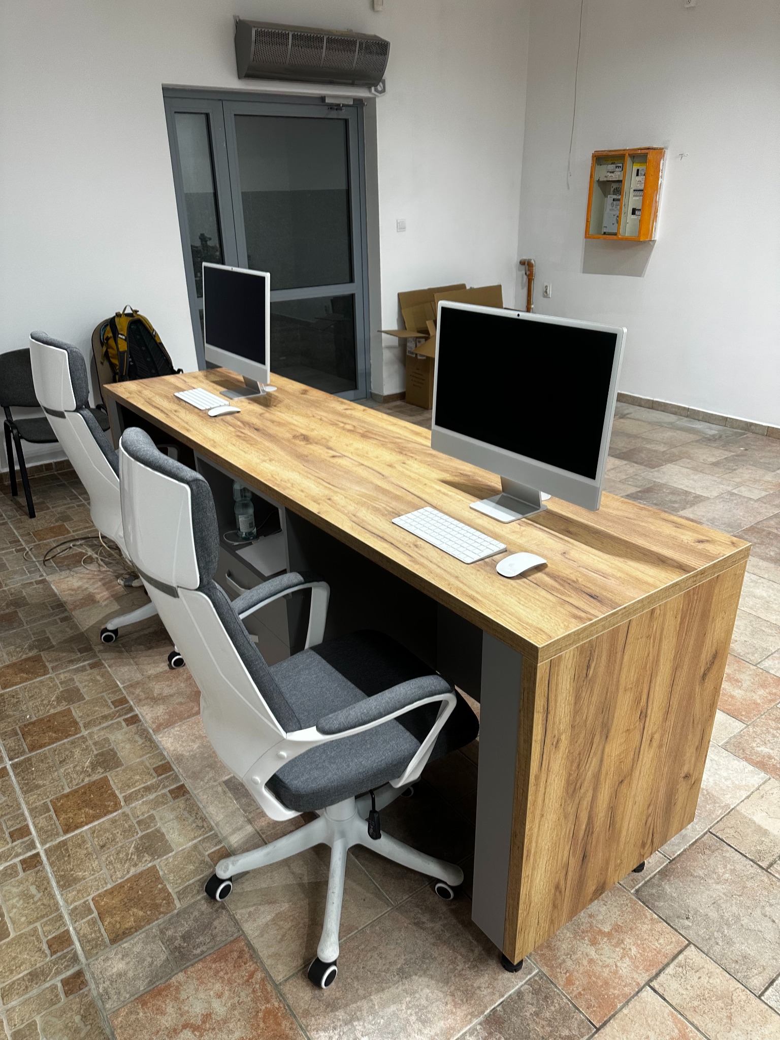 Długi, drewniany stół biurowy z dwoma stanowiskami komputerowymi i ergonomicznymi krzesłami na kafelkowanej podłodze. Widoczne okno i skrzynka elektryczna na ścianie.