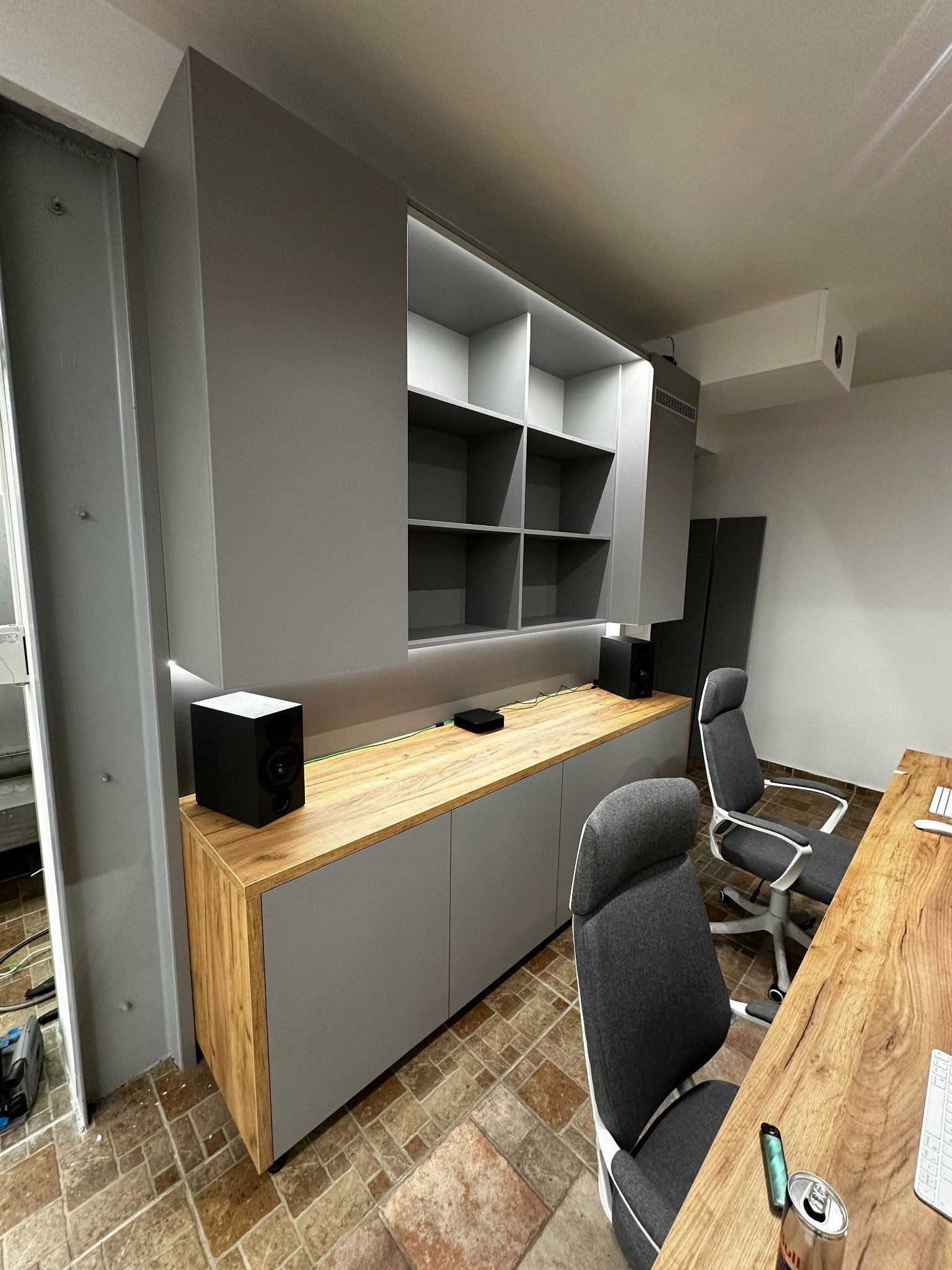 Szara szafa na wymiar z drewnianym blatem i oświetleniem LED, uzupełniona o czarne głośniki. Dwa krzesła biurowe i biurko z blatem z litego drewna w tle.