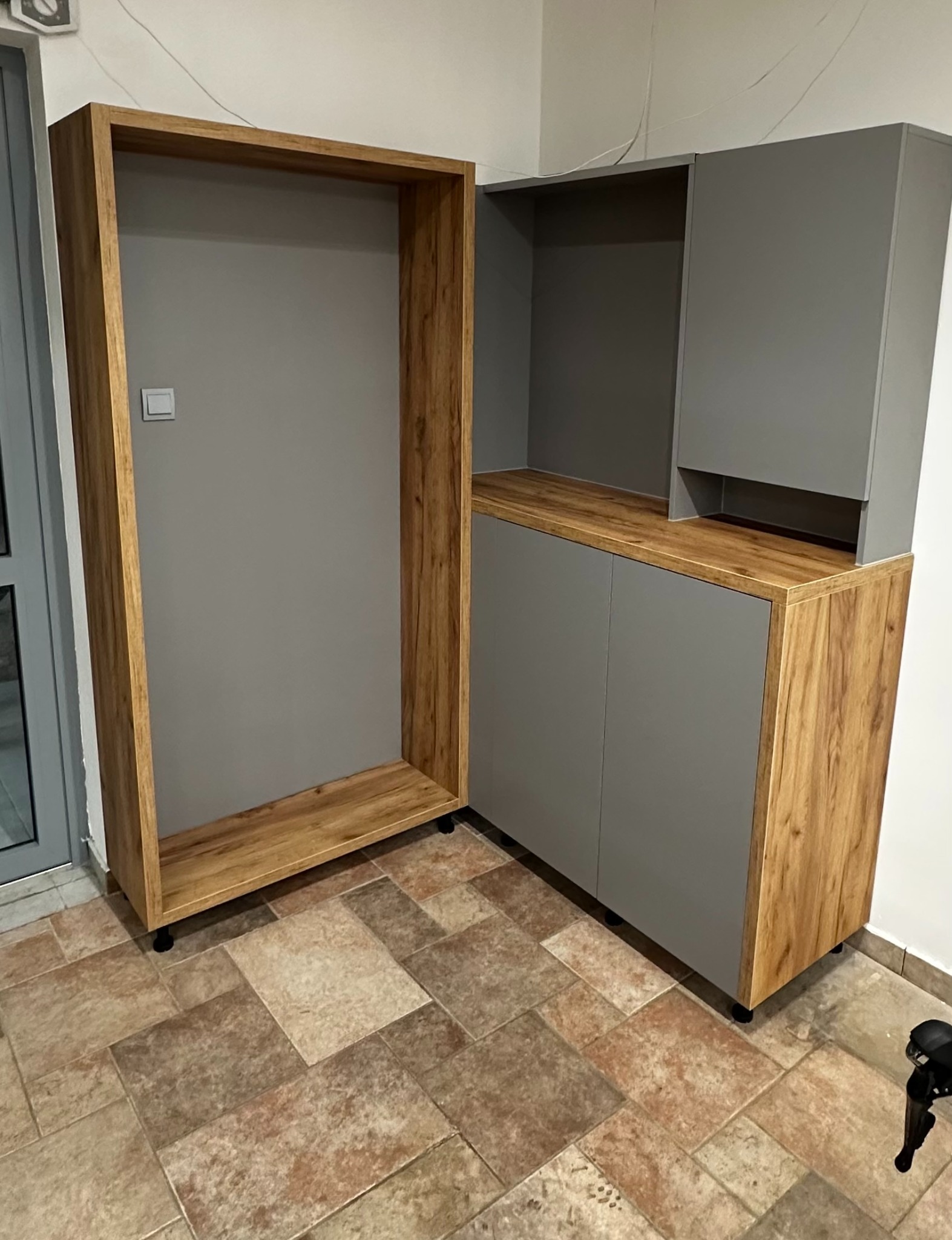Szara szafa z drewnianymi akcentami, widoczna z boku, z otwartą półką na kółkach. Ułożona na brązowej podłodze z płytek.