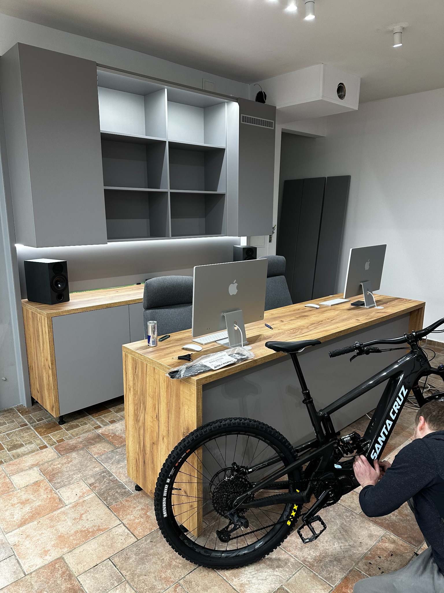 Nowoczesne biuro z szafami na wymiar w odcieniach szarości i drewna, z rowerem Santa Cruz w kadrze. Osoba reguluje przerzutki. Dwa monitory Apple na biurku.