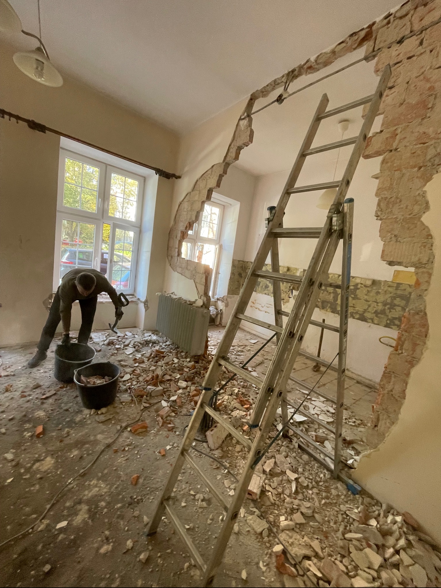 Demontaż ściany z cegły w pomieszczeniu z drabiną i gruzem. Pracownik w trakcie zbierania resztek do wiadra. Widoczne okno z widokiem na zewnątrz.