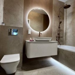 Mar-Motomax Reaktywacja Mariusz Pewniak - Nowoczesna łazienka z okrągłym lustrem LED, białą szafką pod umywalką i beżowymi płytkami. Bateria i deszczownica w kolorze miedzi.