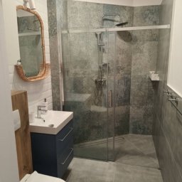 Mar-Motomax Reaktywacja Mariusz Pewniak - Nowoczesna łazienka z kabiną prysznicową typu walk-in, umywalką na szafce w kolorze granatowym i lustrem w ramie z rattanu. Szare płytki imitujące kamień.