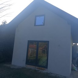 Mar-Motomax Reaktywacja Mariusz Pewniak - Elewacja budynku po szpachlowaniu, widoczne okna z czarnymi ramami i gładka, jasna powierzchnia ściany. Na dole widoczny fragment trawy i resztki materiałów budowlanych.