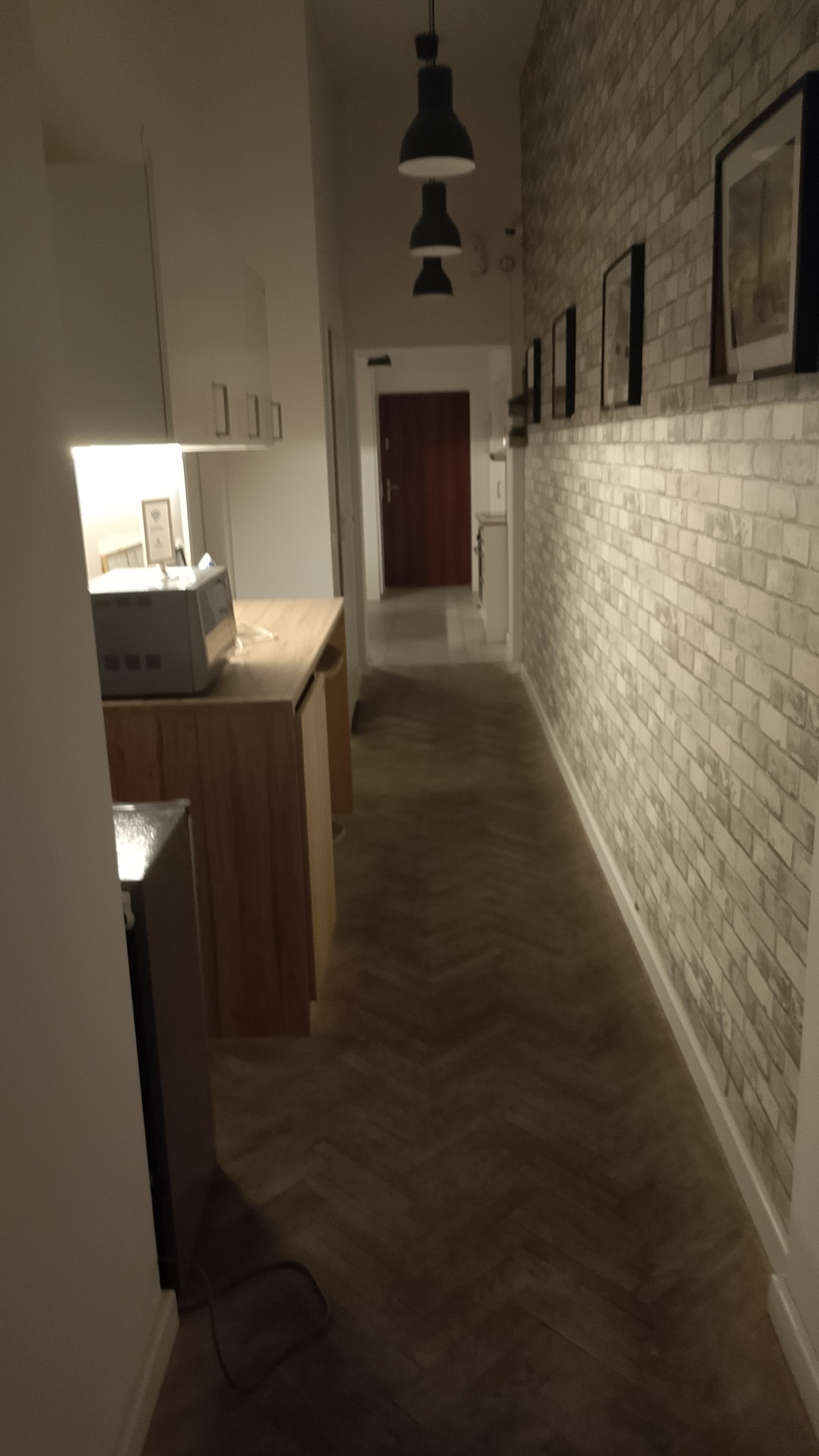 Korytarz z ceglaną ścianą, drewnianą podłogą w jodełkę i nowoczesnym oświetleniem. Widok na kuchnię w tle. Elementy wykończenia wnętrz.