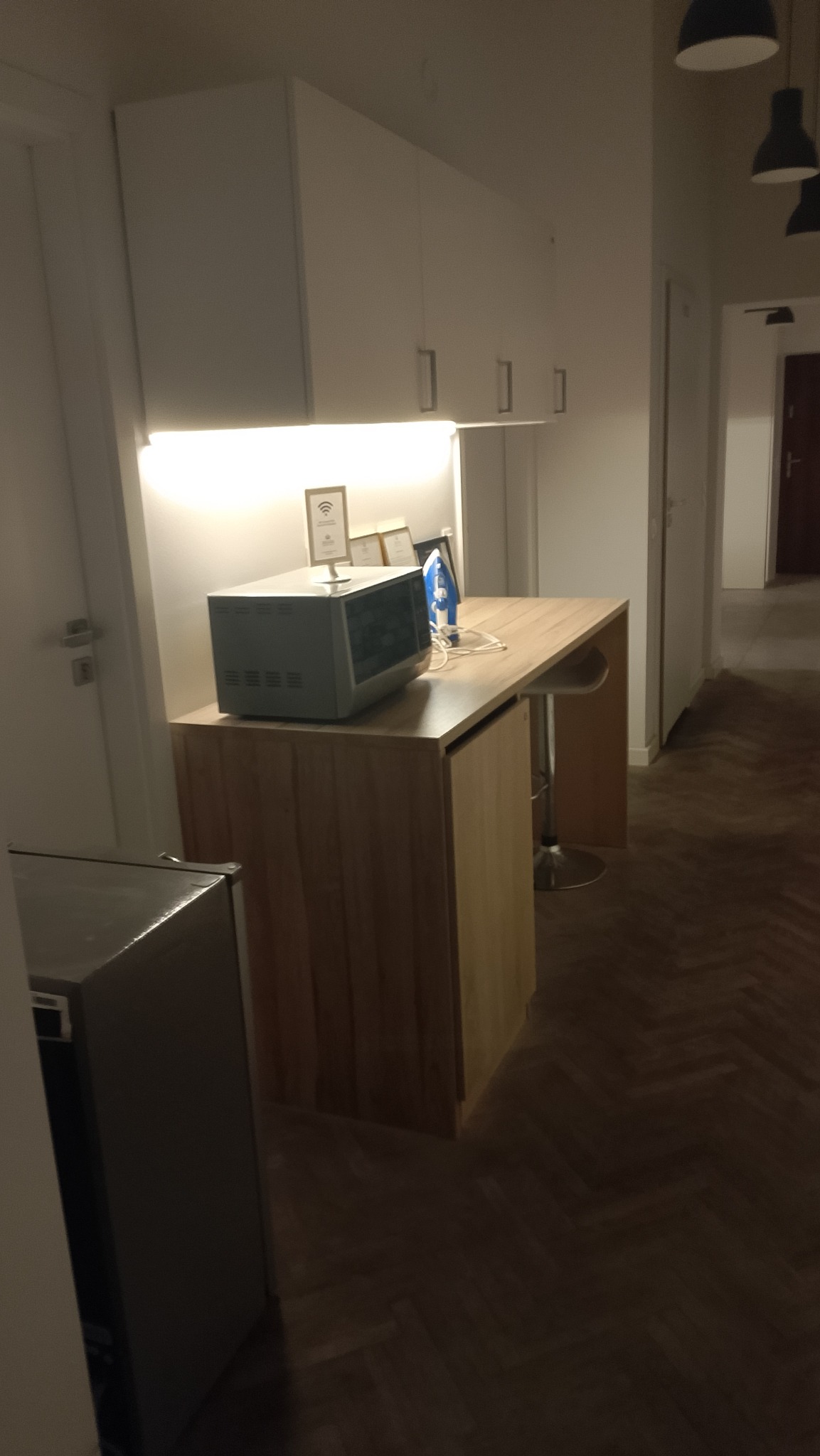 Jasna kuchnia z drewnianym blatem, mikrofalówką i żelazkiem. Białe szafki wiszące i podświetlenie LED. W tle widoczny fragment korytarza i podłoga z parkietu.