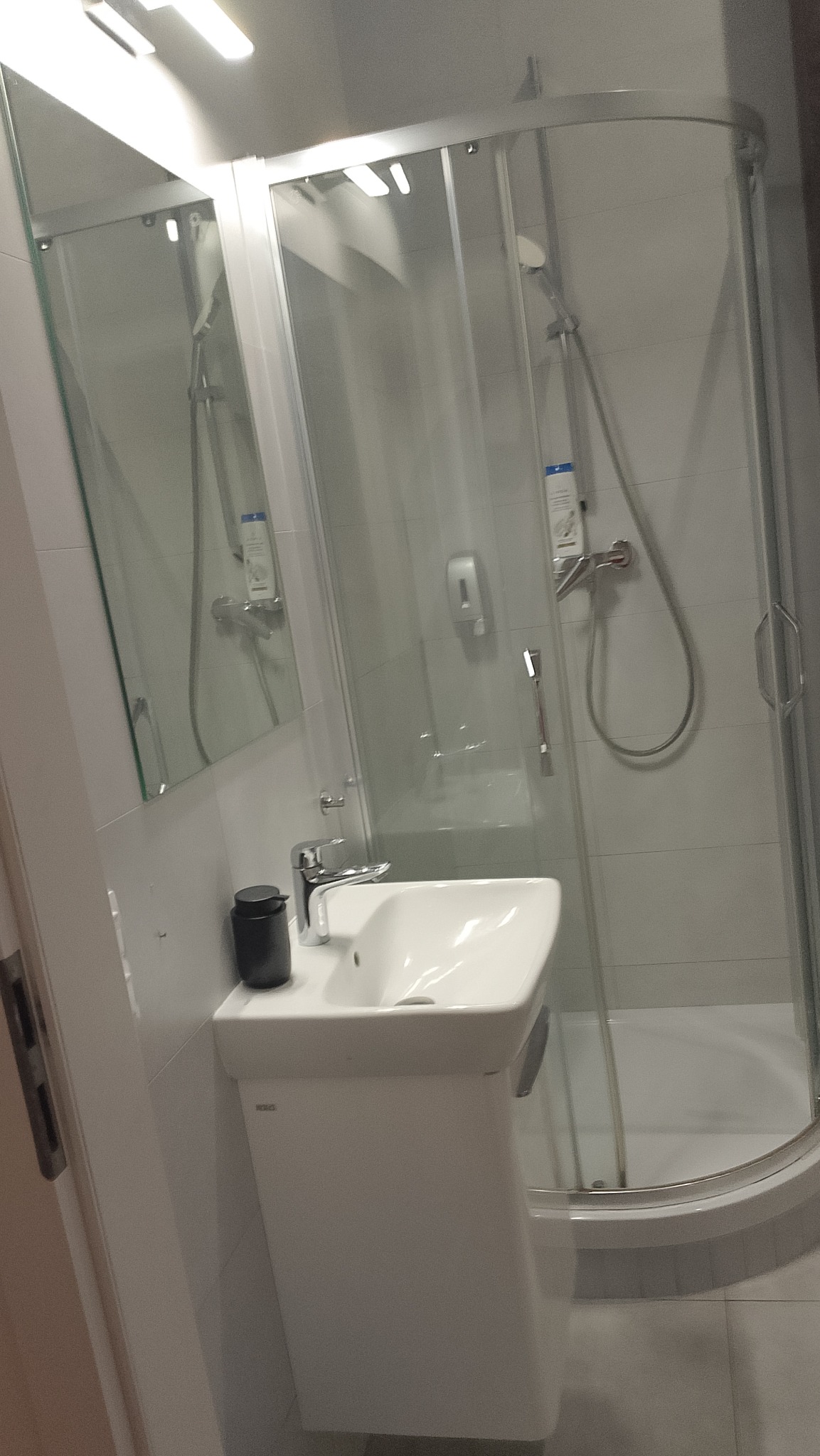 Nowoczesna, jasna łazienka z kabiną prysznicową i białą umywalką na szafce. Widoczne chromowane elementy armatury i minimalistyczny design.