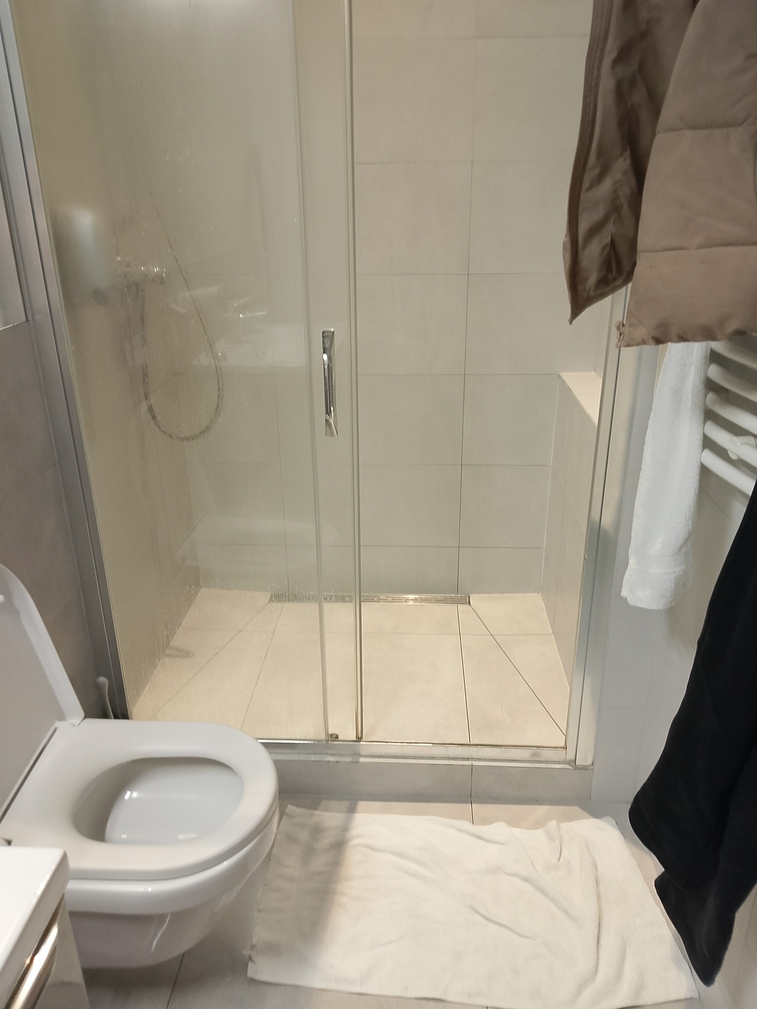 Nowoczesna kabina prysznicowa z toaletą w jasnej łazience, widok z bliska. Na grzejniku wisi ręcznik i kurtka. Minimalistyczny design.