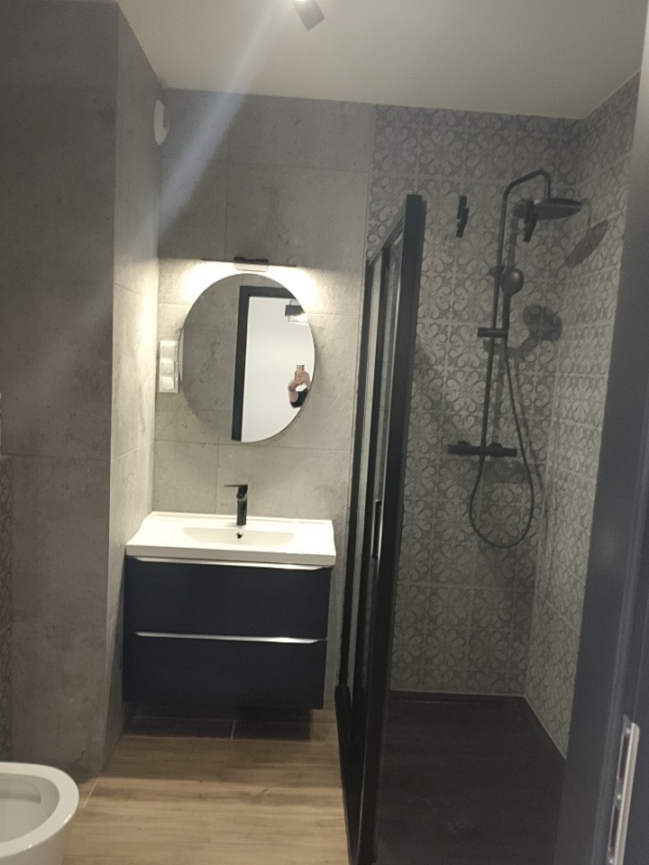 Nowoczesna łazienka z kabiną prysznicową typu walk-in, okrągłym lustrem z podświetleniem i szafką pod umywalką w kolorze granatowym. Minimalistyczny design.