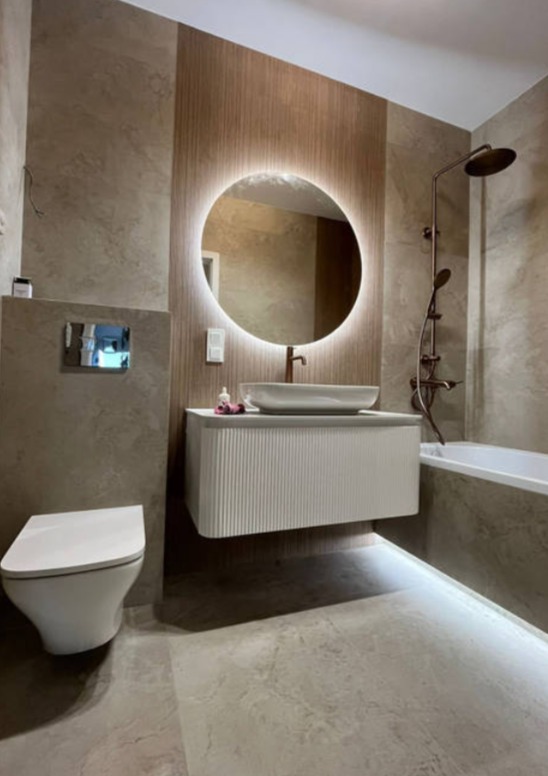 Nowoczesna łazienka z okrągłym lustrem LED, białą szafką pod umywalką i beżowymi płytkami. Bateria i deszczownica w kolorze miedzi.