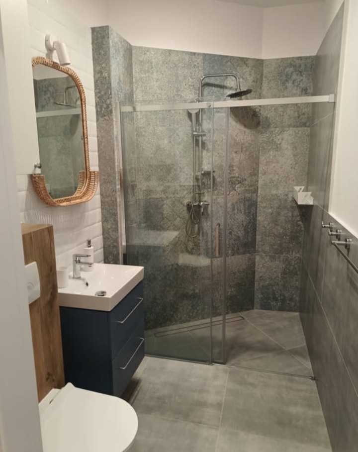 Nowoczesna łazienka z kabiną prysznicową typu walk-in, umywalką na szafce w kolorze granatowym i lustrem w ramie z rattanu. Szare płytki imitujące kamień.