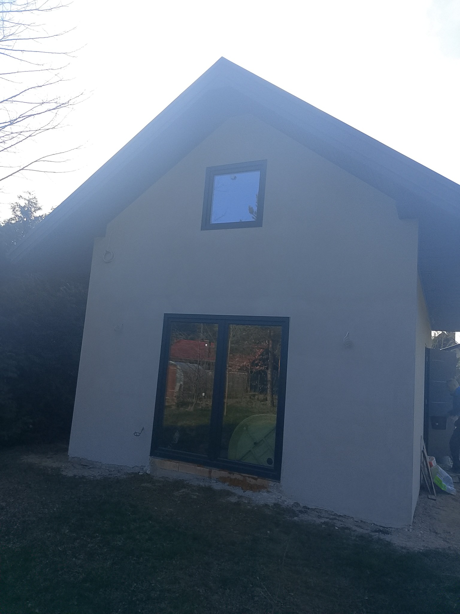 Elewacja budynku po szpachlowaniu, widoczne okna z czarnymi ramami i gładka, jasna powierzchnia ściany. Na dole widoczny fragment trawy i resztki materiałów budowlanych.