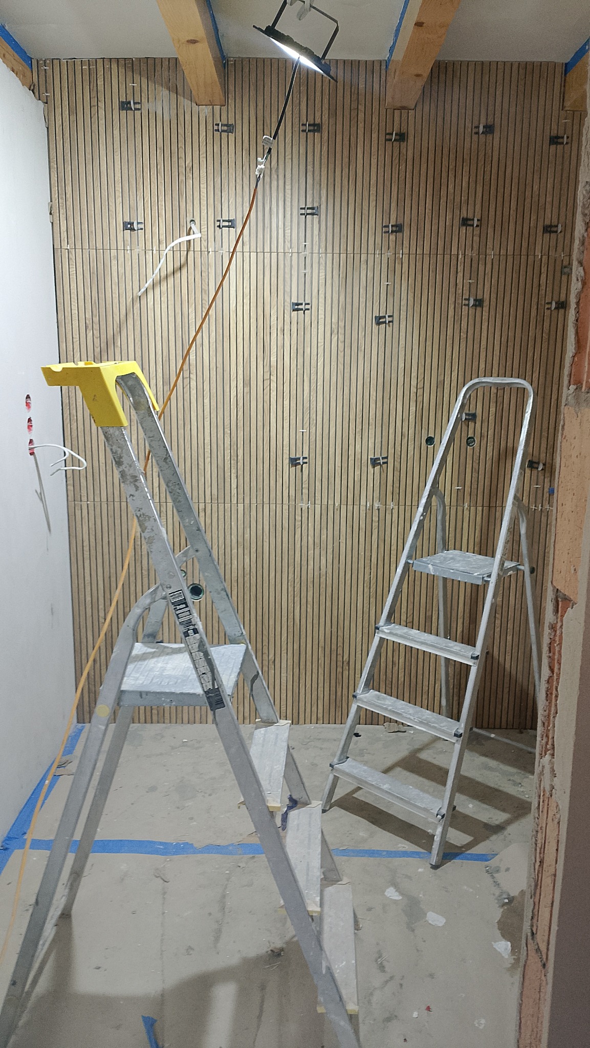 Montaż paneli ściennych z drewnopodobnym wzorem w trakcie realizacji, widoczne drabiny i oświetlenie robocze. Detal wykończenia wnętrza w surowym stanie.