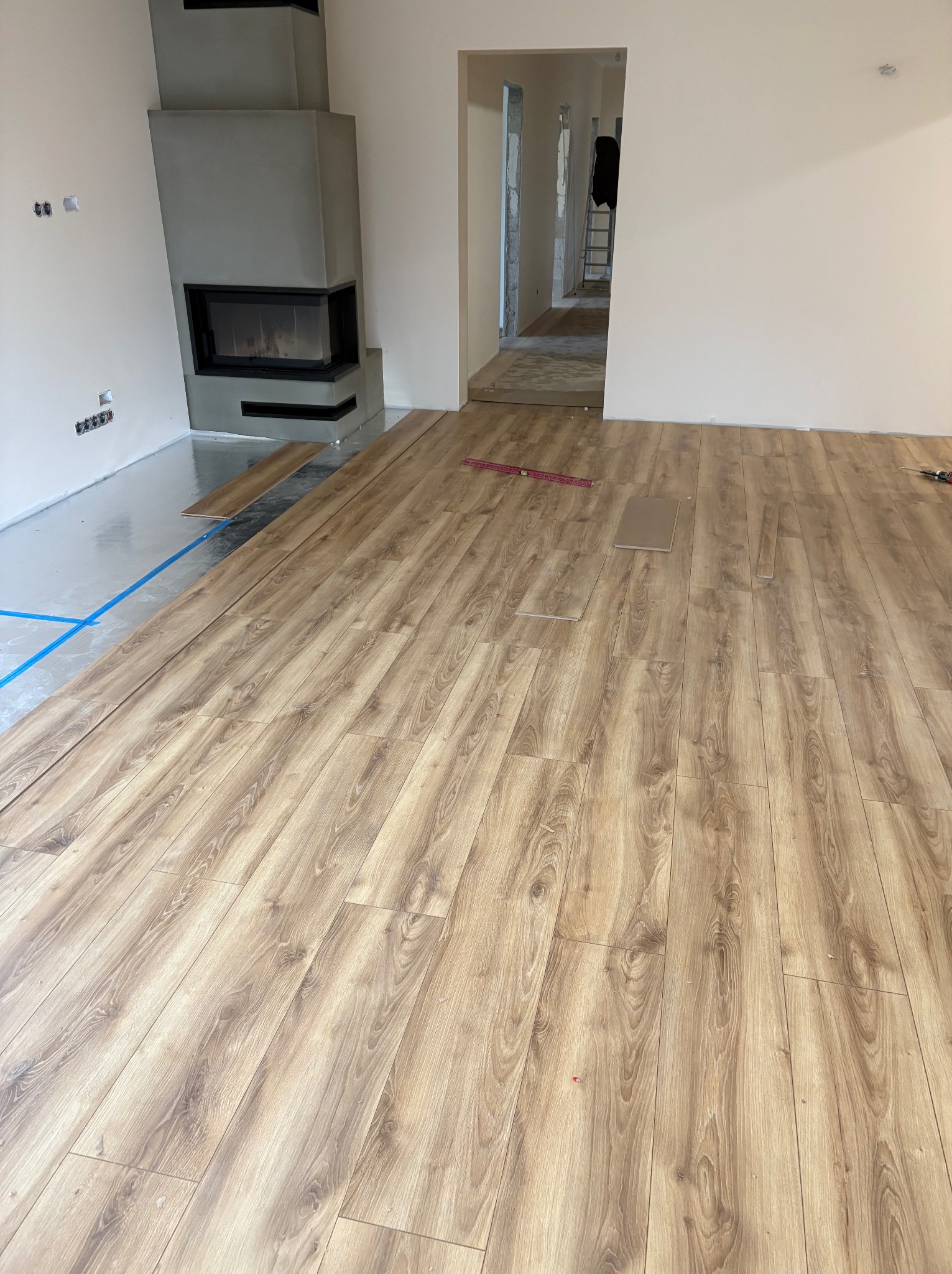 Układanie paneli podłogowych w jasnym wnętrzu z kominkiem. Widoczne narzędzia i poziomica. Częściowo ułożona podłoga z drewnopodobnym wzorem.