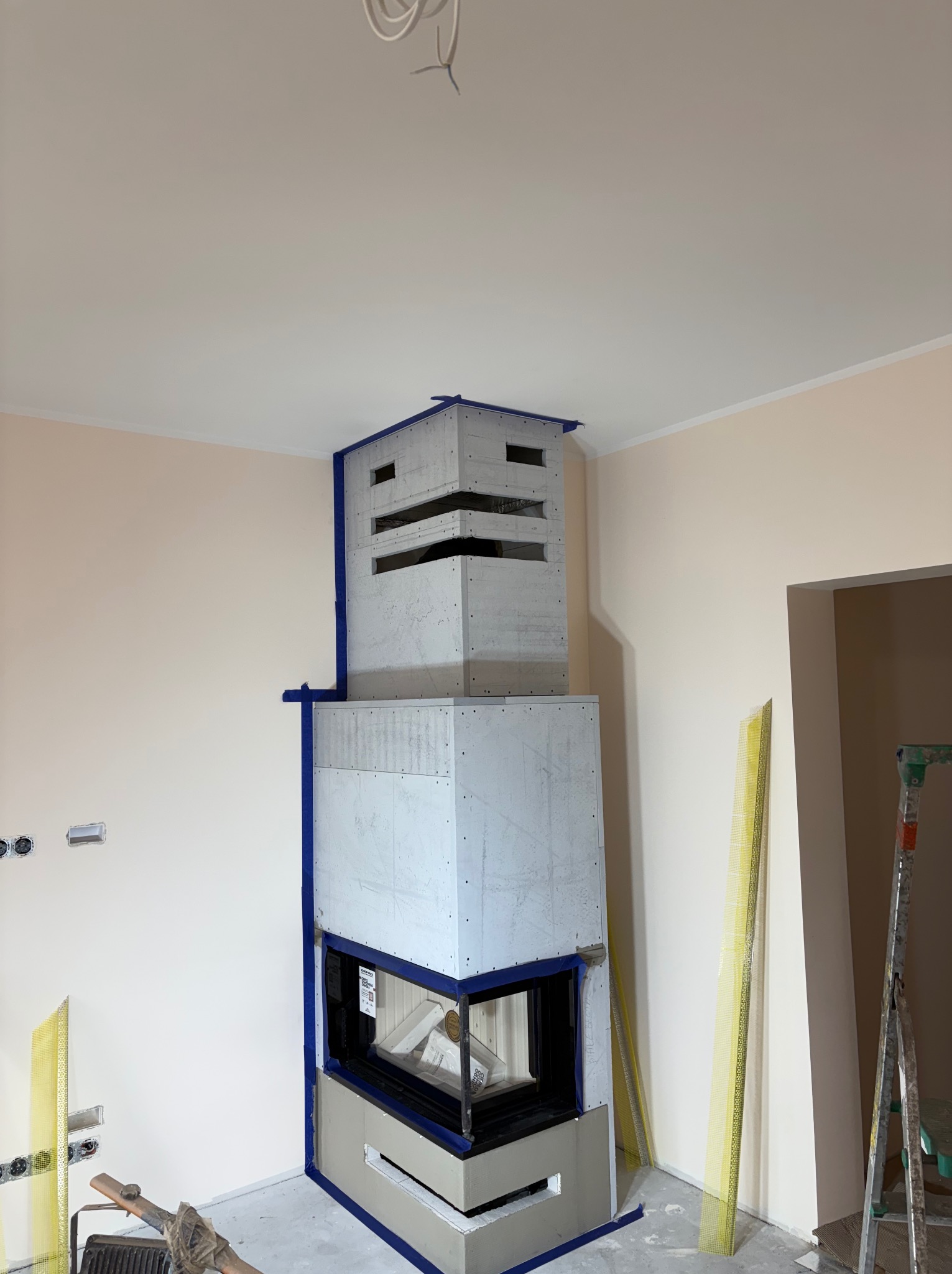 Wnętrze w trakcie wykończenia: obudowa kominka z płyt gipsowych zabezpieczona taśmą, widoczne narzędzia i drabina. Ściany w jasnym odcieniu beżu, sufit biały.