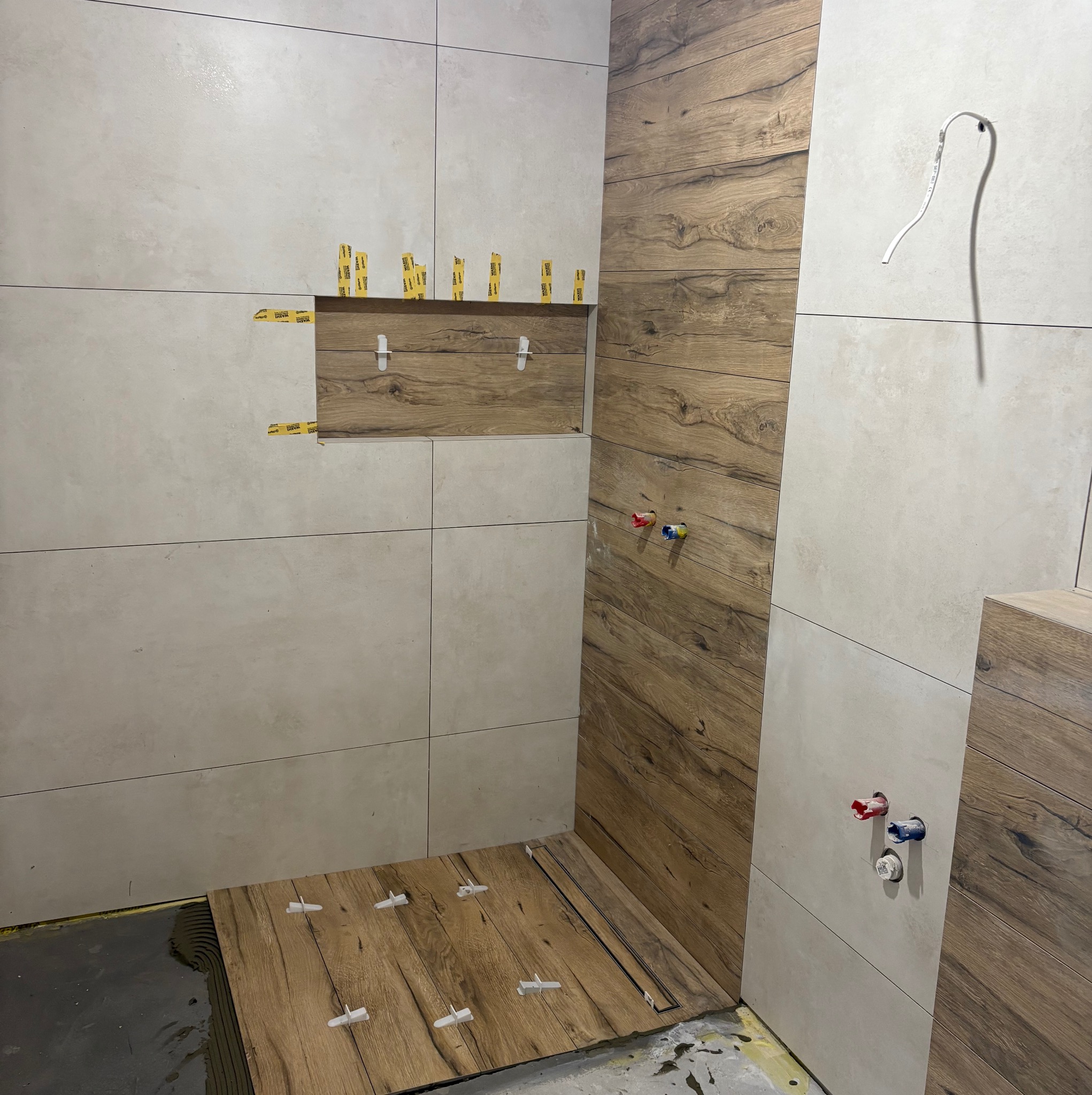 Układanie płytek imitujących drewno i beżowych w łazience. Widoczne krzyżyki dystansowe i taśmy mocujące. Częściowo wykończona ściana i podłoga.