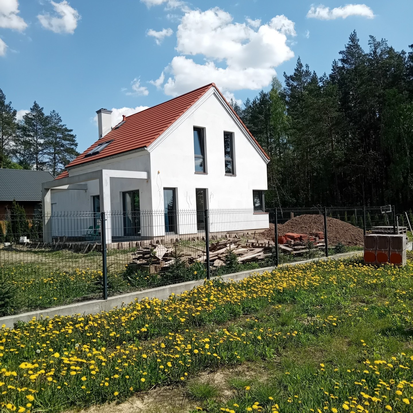 Nowoczesny, biały dom z czerwoną dachówką w otoczeniu zieleni i żółtych kwiatów, ogrodzony czarnym płotem. Widoczne materiały budowlane sugerują etap wykończeniowy.