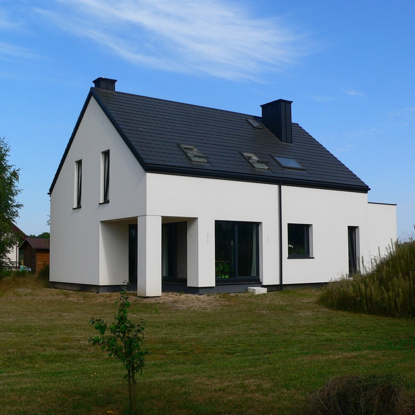 Nowoczesny, biały dom jednorodzinny z ciemną dachówką i minimalistycznym wejściem, otoczony trawnikiem i młodym drzewkiem. Widok z perspektywy ogrodu w słoneczny dzień.