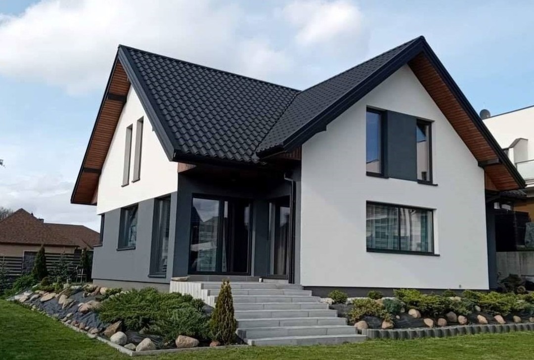 Nowoczesny dom jednorodzinny z ciemną dachówką i jasną elewacją, widok z ogrodu, schody wejściowe, starannie zagospodarowany teren zielony z niską roślinnością.