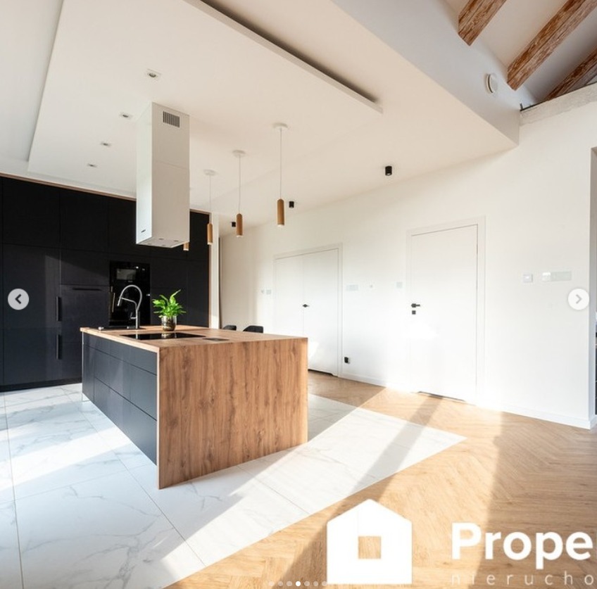 Nowoczesna kuchnia z wyspą: czarne szafki, drewniany blat, białe marmurowe płytki podłogowe i parkiet w jodełkę. Minimalistyczne lampy wiszące i biały okap.