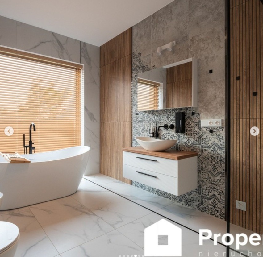 Nowoczesna łazienka z wolnostojącą wanną, umywalką nablatową i drewnianymi akcentami. Marmurowe płytki i geometryczny wzór na ścianie dodają elegancji.