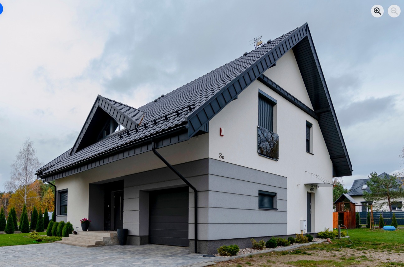 Nowoczesny dom jednorodzinny z garażem, wykończony w bieli i szarości, z ciemną dachówką i elementami stolarki. Widok z zewnątrz, w tle zieleń i pochmurne niebo.