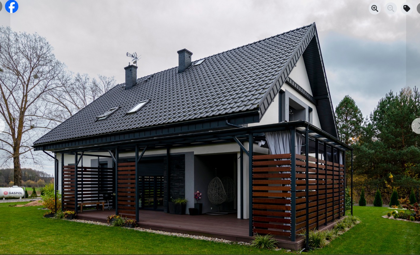 Nowoczesny dom jednorodzinny z zadaszonym tarasem i drewnianą zabudową. Czarny dach, biała elewacja, zadbany trawnik. Widok z zewnątrz, w tle drzewa i zbiornik na gaz.