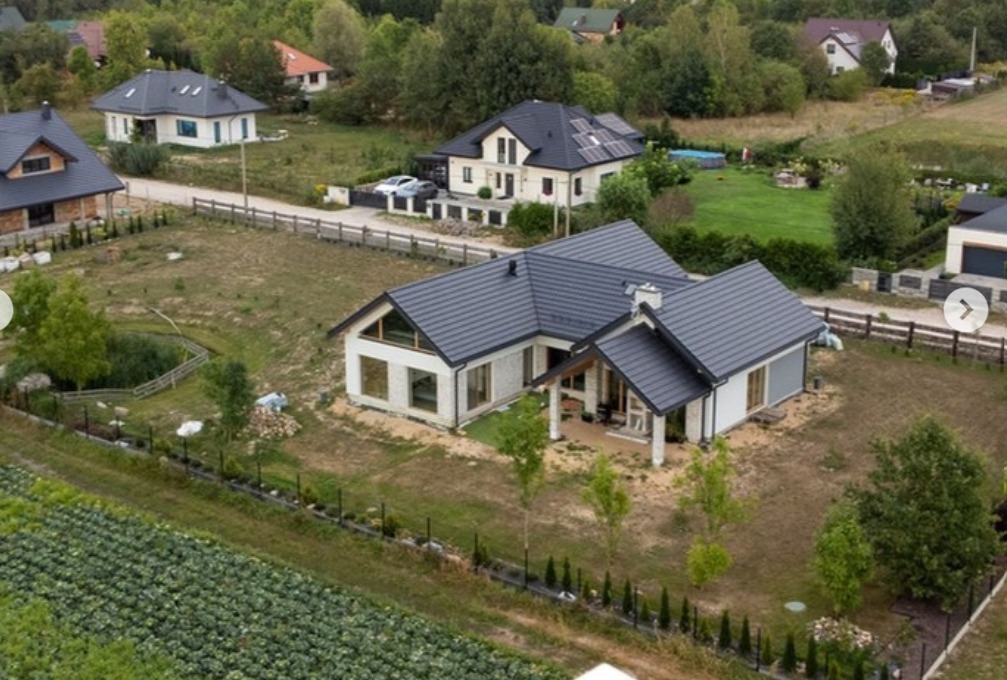 Widok z lotu ptaka na nowoczesny dom jednorodzinny z ciemnym dachem, jasną elewacją i dużymi oknami, otoczony zielenią i innymi zabudowaniami.