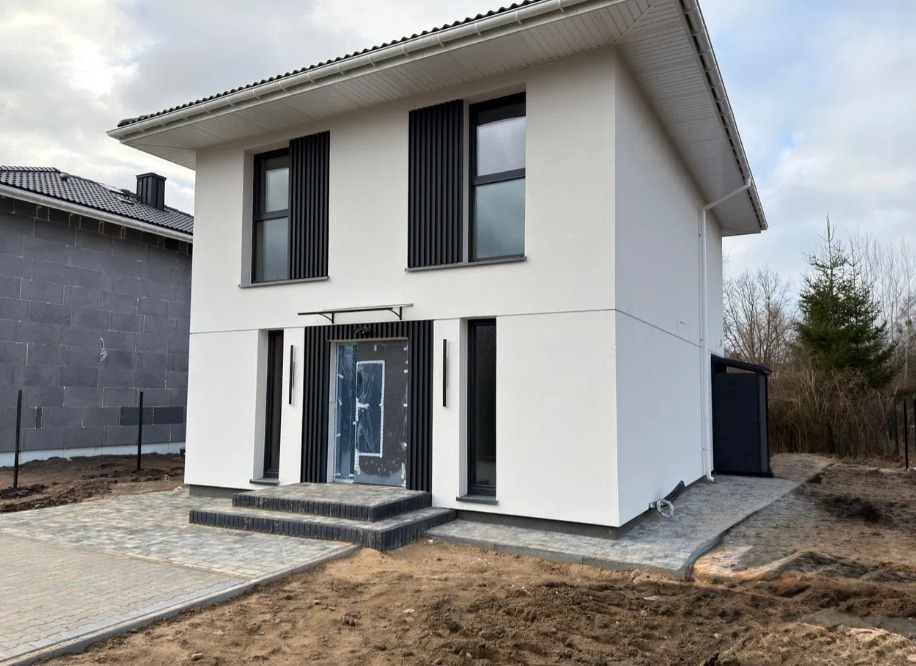 Nowoczesny, dwukondygnacyjny dom z białymi ścianami i czarnymi akcentami, takimi jak żaluzje okienne i obramowania. Wejście zadaszone, schody z kostki brukowej.