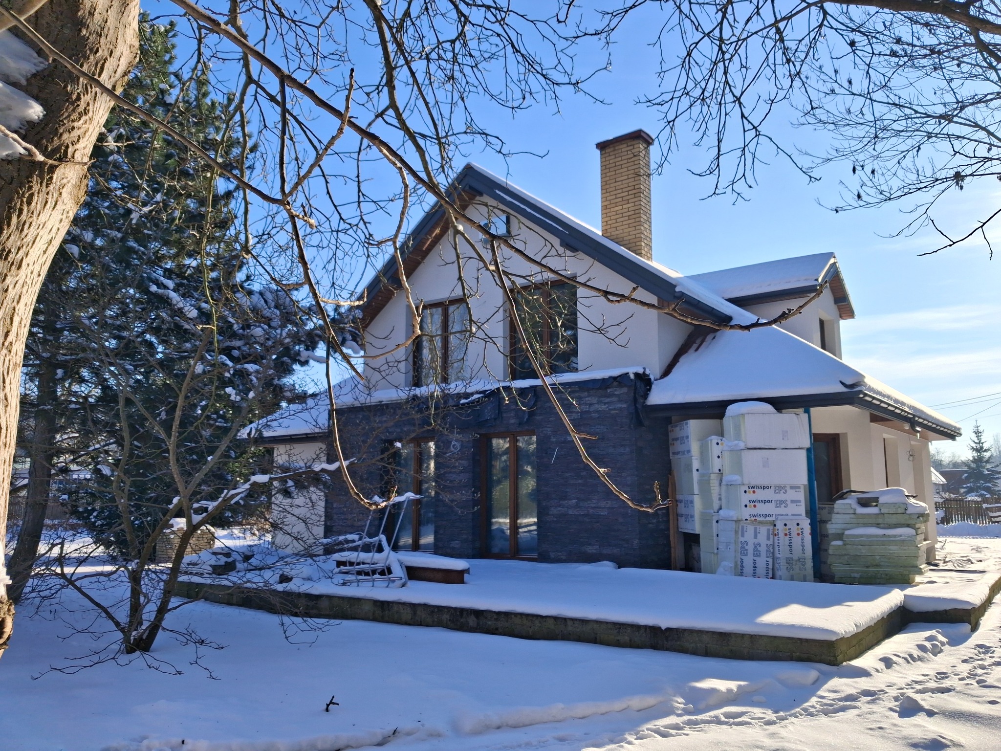 Dom jednorodzinny w trakcie przebudowy zimą, z widocznym ociepleniem i elementami wykończeniowymi. Śnieg pokrywa teren wokół budynku i materiały budowlane.