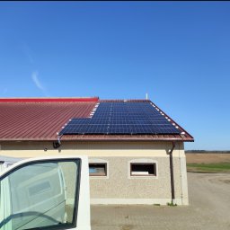 Volt Master Bartosz Cybulski - Panele słoneczne na dachu budynku z czerwoną dachówką, widok z boku z częściowo widocznym białym samochodem dostawczym. Błękitne niebo w tle.