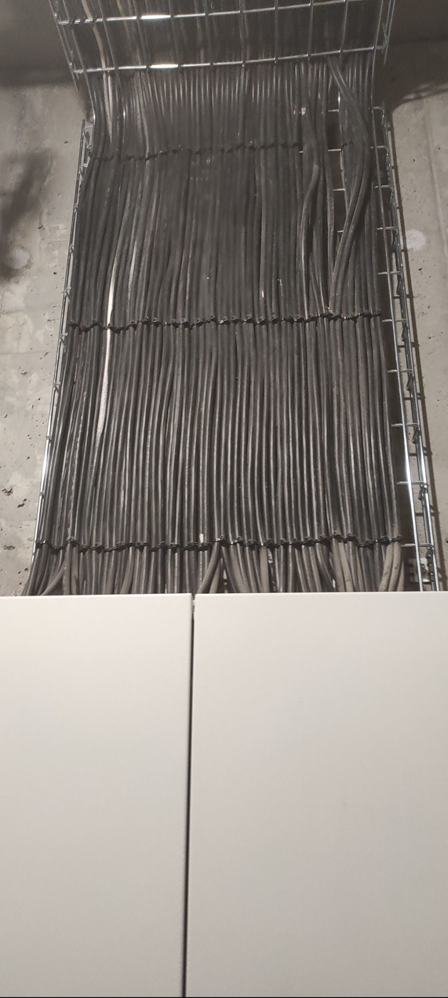 Uporządkowane kable w metalowej korycie, widoczne szare przewody ułożone równolegle, z białym panelem na dole kadru. Instalacja w surowym otoczeniu.