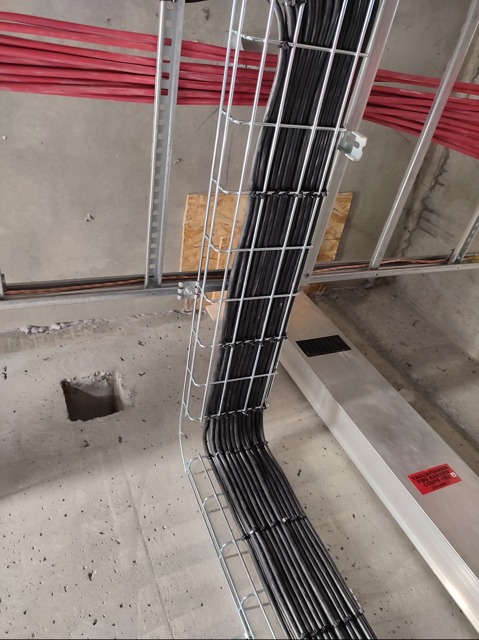 Kable w metalowej korycie przymocowane do sufitu, widoczne czerwone rury. Instalacja kablowa w budynku w trakcie realizacji. Widok z dołu, surowy beton.