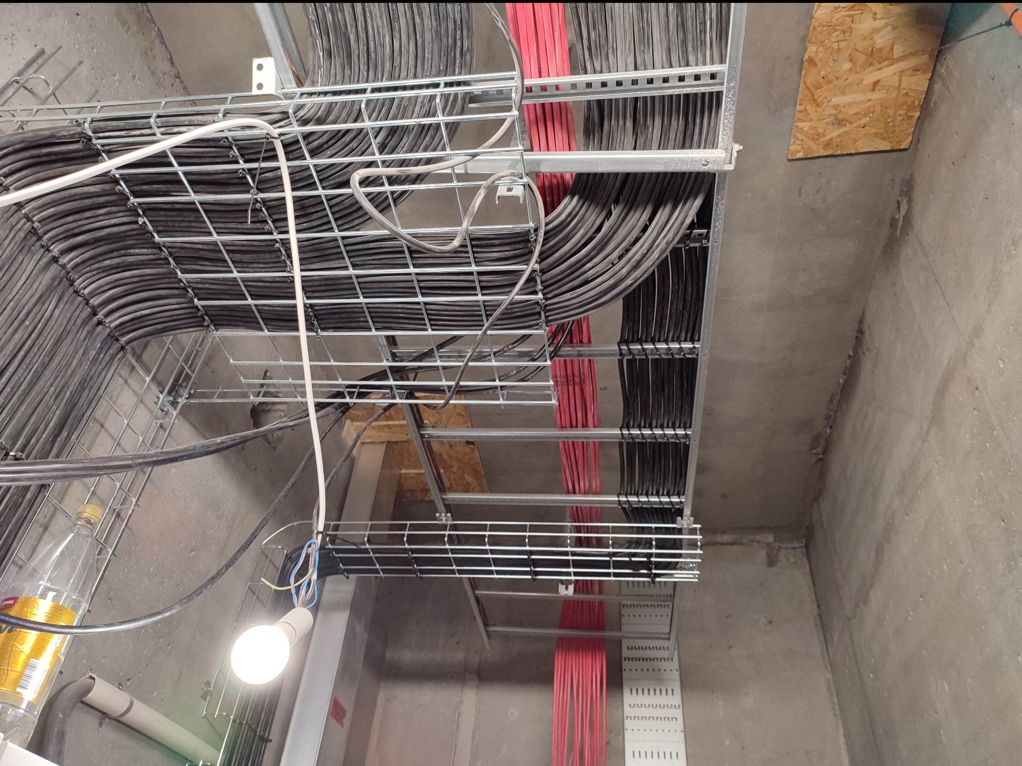 Kable elektryczne ułożone w metalowych korytkach pod sufitem, widoczne czarne i czerwone przewody. Oświetlenie tymczasowe z żarówką. Surowy betonowy sufit i ściany.